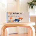 A box of the Magblox Twin Car Set, a STEAM educational toy from Magblox featuring car illustrations, geometric shapes, and a racing flag, is positioned on a round wooden stool in a bright room with sunlight streaming through. The box indicates it contains two pieces and is suitable for ages 1-99, encouraging creative play.