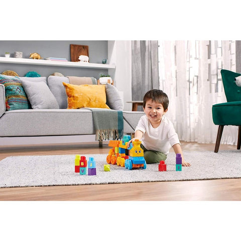 In a well-lit living room flooded with natural light, a young child sits on a carpeted floor, smiling as they play with colorful Mega Bloks and the Mega Bloks ABC Musical Train. A sofa adorned with cushions and a bookshelf filled with decorative items can be seen in the background, creating the perfect setting for an alphabet adventure.