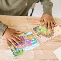 An individual in a green long-sleeve shirt is seen on a wooden table, assembling two vibrant wooden puzzles from the "Melissa & Doug Dinosaurs Puzzles in a Box" set by Melissa & Doug. Using both hands, they piece together illustrations featuring tropical foliage and an animated purple dinosaur.