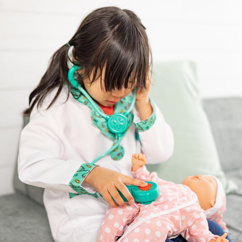 A young child dressed in the Melissa & Doug Doctor Role Play Costume Set pretends to examine a doll using a toy stethoscope. The child wears the white coat and has the green toy stethoscope with sound effects from the set around their neck, while the doll is dressed in a pink outfit with white polka dots.