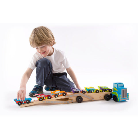 A child kneels on the ground, playing with the Melissa & Doug Mega Race-Car Carrier. The colorful wooden set features an empty truck with a detachable trailer, and six small race cars in different colors positioned on the trailer. This engaging activity helps develop motor skills. The child is wearing a white t-shirt and dark pants.