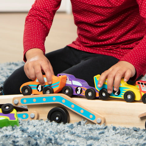 A child wearing a red polka dot shirt plays with colorful wooden toy race cars on a wooden ramp, honing their motor skills. The toys include cars numbered 4, 3, and 2, each featuring vibrant designs. Nearby on the soft blue carpet lies the Melissa & Doug Mega Race-Car Carrier by Melissa & Doug.