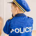 A young child, seen from behind, is dressed in the Melissa & Doug Police Officer Costume Role Play Set. The costume features a blue police officer uniform and cap with yellow stripes, with "POLICE" printed in white letters on the back. The child appears to be holding a walkie-talkie with sound effects close to their mouth.