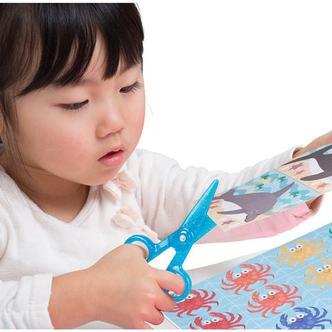 A young child with dark hair is deeply concentrating while using blue safety scissors from the Melissa & Doug Sea Life Scissor Skills activity pad to cut a piece of paper featuring cartoon sea animals like sharks and crabs. The child, wearing a white long-sleeve shirt, appears thoroughly engaged in the project, honing their scissor skills with the help of this fun and educational product from Melissa & Doug.