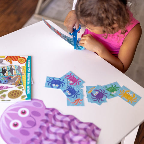 A young child practices scissor skills using Melissa & Doug Sea Life Scissor Skills with the blue scissors, cutting out a colorful octopus-shaped paper from the Ocean-themed activity pad by Melissa & Doug on a white table. Nearby are multiple cut-out pieces of marine shapes and a booklet with more ocean animal illustrations.