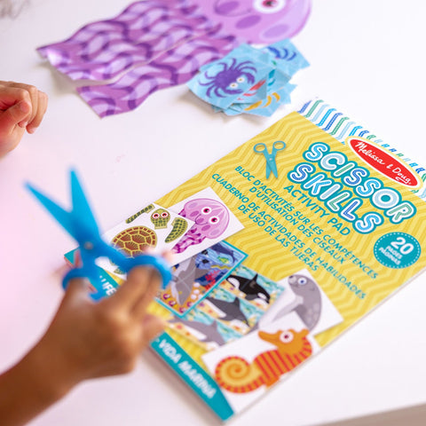 A child uses blue scissors to cut paper on a table. In front of them is a "Melissa & Doug Sea Life Scissor Skills" pad with illustrations of ocean creatures. The table has cut-out images of sea animals, and the child holds one of the paper-cutting projects in hand.
