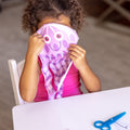 A young child with curly hair sits at a white table, holding a pink and purple jellyfish paper craft up to their face. The jellyfish has a smiley face and tentacles. On the table is a pair of blue scissors from the Melissa & Doug Sea Life Scissor Skills set, perfect for enhancing scissor skills through fun ocean-themed activity pad projects by Melissa & Doug.