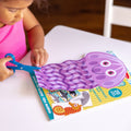 A child in a pink shirt is developing their scissor skills by cutting out a purple, smiling jellyfish from the colorful Melissa & Doug Sea Life Scissor Skills activity pad using a pair of blue scissors. The activity book from Melissa & Doug also features a sea horse and other marine-themed illustrations.