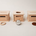 Three wooden boxes are aligned horizontally against a plain background. The center box, a Navy Baby Posting Box with Parts by Navy Baby, has a circular opening on top, with a shiny metal sphere placed directly in front of it. There is a wooden ring to the left and a large button to the right of the boxes, offering open-ended options for play.