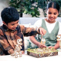 Two children sit at a table playing Orchard Toys Dinosaur Dig Game by Orchard Toys. The boy on the left, wearing a plaid shirt, is holding a small wooden dinosaur piece, while the girl on the right, in a green dress, smiles and looks on. The game helps with matching and memory skills as well as dexterity development. There is greenery in the background.