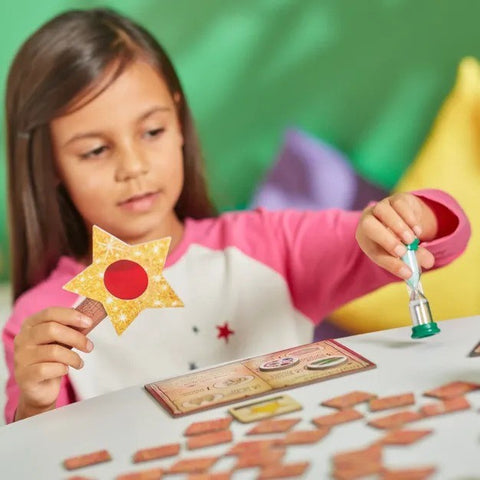 A young girl with long brown hair, wearing a pink and white shirt, is deeply engrossed in playing Orchard Toys' Magic Spelling game. She holds a star-shaped piece in one hand while turning over a small hourglass timer with the other, creating an enchanting scene reminiscent of a spell book. The table is scattered with various pieces from the Orchard Toys game set.