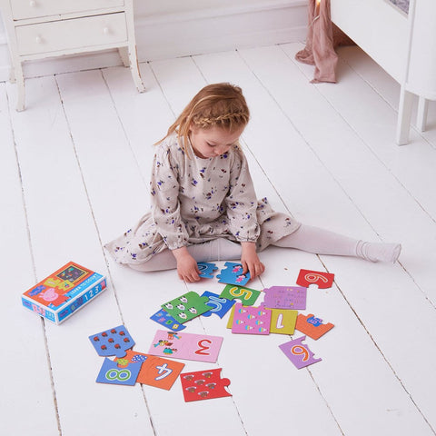 A young child sits on a white wooden floor, wearing a light-colored dress and white tights. She is playing with colorful puzzle pieces featuring numbers and images of objects. Nearby is a blue box labeled "Peppa Pig Learn to Count Puzzle," an educational game by Holdson that helps kids match numbers.