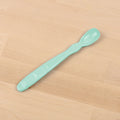 The REPLAY Re-Play Infant Spoon, in a light green color, is an elongated baby spoon crafted from recycled material. It features a small, rounded scoop on one end and a flat handle with textured grips for easy handling. Perfect for sustainable tableware collections, it rests elegantly on a wooden surface.