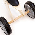 Close-up of a wooden, three-wheeled Runna Mini - Gumtree trike by RUNNA, focusing on the two rear black tires with grey rims. The frame is made from light-colored wood, and part of the black seat is visible. Perfect for toddlers learning balance, the background is plain white.