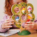 A child with curly red hair plays with the Smart Games 5 Little Birds wooden puzzle by Smart Games, which is shaped like a tree and features colorful bird figures perched on its branches. The child arranges the birds, enhancing their cognitive skills, while an open activity book lies on the table beside them.