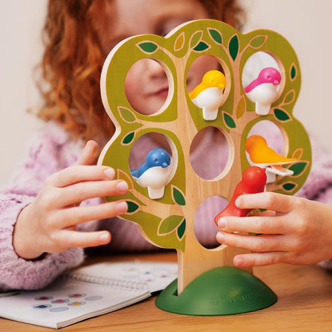 A child with curly red hair plays with the Smart Games 5 Little Birds wooden puzzle by Smart Games, which is shaped like a tree and features colorful bird figures perched on its branches. The child arranges the birds, enhancing their cognitive skills, while an open activity book lies on the table beside them.