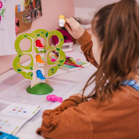 A child plays with the vibrant Smart Games 5 Little Birds wooden tree puzzle by Smart Games, attaching various bird figures. The child is placing a white and orange bird at the top of the tree. Scattered across the table are art supplies and a partially opened book or manual, enhancing their cognitive skills as they engage in play.