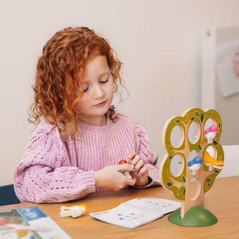 A young child with curly red hair wearing a pink sweater sits at a table, assembling the Smart Games 5 Little Birds puzzle with its colorful pieces. They are examining the toy and holding a piece in their hand, with a booklet open in front of them, enhancing their cognitive skills in this engaging activity from Smart Games.