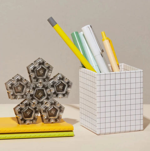 A white grid-patterned pencil holder containing yellow, green, gray, and white pens stands on a surface. Beside it is a set of Speks Slate Magnetic Pentagons - Set of 12 in silver hexagonal shape resembling a mechanical puzzle resting on a yellow notebook. The background is a light neutral color.