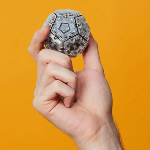 A hand holds a transparent Speks Slate Magnetic Pentagons - Set of 12^ against an orange background. The puzzle ball, resembling a geode, has a multifaceted design with magnetic pentagons and small hexagonal panels linked together.