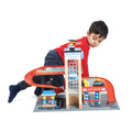 A young boy in a red shirt and dark pants is playing with the Tender Leaf Blue Bird Service Station, a toy parking garage set. The set features a building labeled "Bluebird," a car wash, a gas station, and a red ramp. Engrossed in imaginative play for children, he positions himself next to the wooden cars playset.