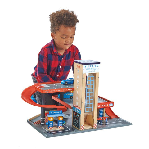 A young child with curly hair, wearing a red and navy plaid shirt, plays with the Tender Leaf Blue Bird Service Station. This imaginative play set for children comes from the brand Tender Leaf and features ramps, a car wash, a gasoline station, and a parking lot filled with wooden cars in blue and red.