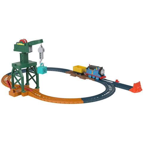 The Fisher-Price Thomas & Friends Motorized Engine - Cranky The Crane playset features a blue toy train on a circular motorized track set with an orange section. There is a tall green crane with a gray hook lifting a green crate. The train, resembling characters from Thomas & Friends, is pulling a yellow cargo car. An orange rock is situated beside the track.