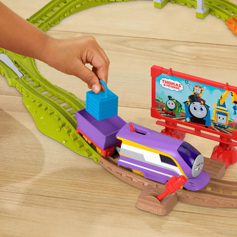 A child's hand places a blue block onto the Fisher-Price Thomas & Friends Motorized Engine - Round & Round Kana toy train on a track. In the background, there is a colorful billboard featuring characters from "Thomas & Friends," and an intricate green and brown motorized train set layout on a light wood surface.