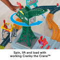 A child's hand is operating a green crane toy labeled "Cranky the Crane™." The crane, part of the Thomas & Friends Talking Cranky Delivery Train Set by Fisher-Price, is positioned on a track set featuring various connected rails and accessories. It is lifting a red load with its hook. Text below reads: “Spin, lift and load with working Cranky the Crane™.”