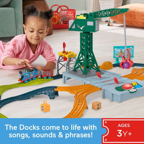 A young child is playing with the Fisher-Price Thomas & Friends Talking Cranky Delivery Train Set, complete with a crane, tracks, and various accessories. This vibrant set bursts with colors and brings The Docks to life with songs, sounds, and phrases. Suitable for ages 3+, as indicated in the text on the image.