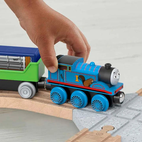 A child’s hand is seen playing with a blue Thomas & Friends Wooden Railway train featuring the number "1" and an image of a beaver on its side. The Fisher-Price train is connected to a green and blue freight car loaded with logs, all situated on the wooden track included in the Thomas & Friends Wooden Railway Figure 8 Track Pack, making it ideal for preschool kids.