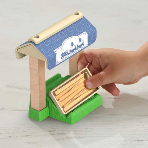 A child's hand places a small wooden crate into the toy structure resembling a train platform with a roof. The Thomas & Friends Wooden Railway Figure 8 Track Pack by Fisher-Price features a blue roof with a white train logo and is supported by two wooden posts on a green base—perfect for preschool kids.