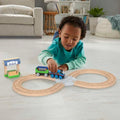 A young child wearing a teal shirt and dark green pants is playing with the Fisher-Price Thomas & Friends Wooden Railway Figure 8 Track Pack on a light-colored floor in a cozy, well-lit living room. The wood train set includes tracks arranged in a figure-eight pattern and a small station, perfect for preschool kids.