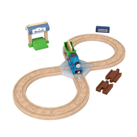 The Fisher-Price Thomas & Friends Wooden Railway Figure 8 Track Pack is shown, featuring a charming wood train set with an oval-shaped track layout. On the track is a blue train with a smiling face, reminiscent of Thomas & Friends. Additional pieces include a green and cream station, a blue sign, and two small brown track connectors—perfect for preschool kids.