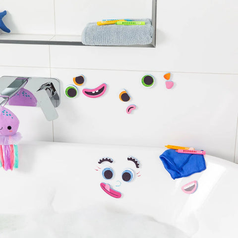 A white bathroom wall above a bathtub is adorned with colorful foam facial features, including eyes, mouths, and noses from the Tiger Tribe Bath Faces set by TIGER TRIBE, fostering creative play. A washcloth and a toothbrush rest on the edge of the tub, with a toy octopus hanging nearby.