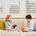 Two young children are sitting on a light-colored sofa, playing the Tiger Tribe Crazy 8s + Go Fish Card Game Set. The girl with glasses and a pink headband holds several cards from the illustrated game set by TIGER TRIBE and smiles. The boy across from her, in a light grey shirt and shorts, is also smiling. Behind them are windows with white shutters.