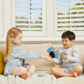 Two young children sit on a beige couch, engrossed in the Tiger Tribe Crazy 8s + Go Fish Card Game Set by TIGER TRIBE. The girl on the left, with blonde hair and glasses, wears a light blue dress and pink leggings. The boy on the right has black hair and is dressed in a grey long-sleeve shirt and light shorts.