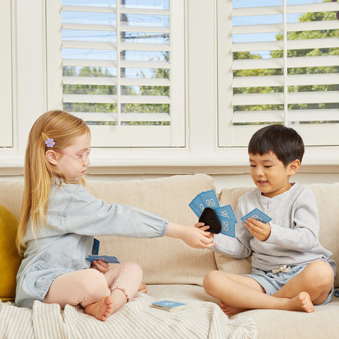 Two young children sit on a beige couch, engrossed in the Tiger Tribe Crazy 8s + Go Fish Card Game Set by TIGER TRIBE. The girl on the left, with blonde hair and glasses, wears a light blue dress and pink leggings. The boy on the right has black hair and is dressed in a grey long-sleeve shirt and light shorts.