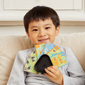 A young child with short dark hair smiles and holds a fan of colorful cards from the Tiger Tribe Crazy 8s + Go Fish Card Game Set by TIGER TRIBE, seated on a light-colored sofa. The child is wearing a light gray outfit, and the background features a white window frame.