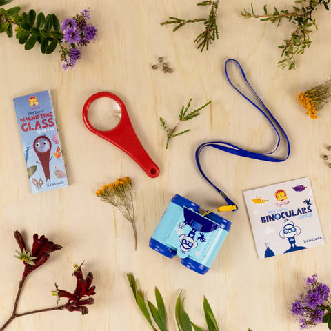 A wooden surface showcases the Tiger Tribe Explorer Magnifying Glass children's adventure set, perfect for nature exploration. This TIGER TRIBE set includes a red magnifying glass, blue binoculars adorned with a cartoon character, an activity booklet for the binoculars, a magnifying glass box, and various scattered flowers and leaves.