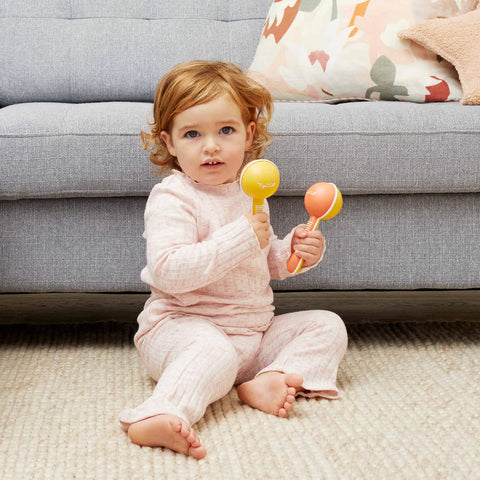 A young child with curly hair sits on a cream-colored carpet in front of a grey sofa, holding two TIGER TRIBE Tiger Tribe Maracas—one yellow and one orange. The child, dressed in a light pink, long-sleeved outfit made from sustainable bio plastic, gazes towards the camera. A pillow with a colorful pattern is on the sofa.