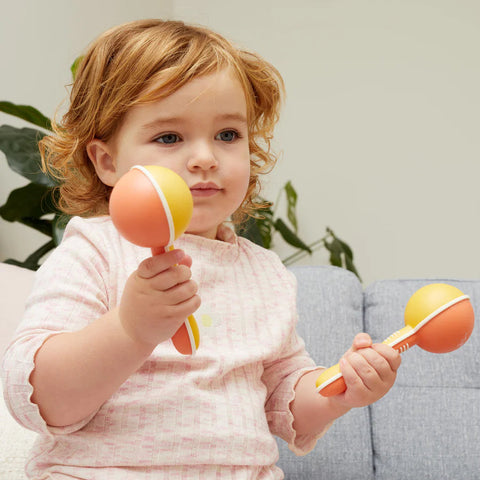 A young child with light brown hair, wearing a light pink sweater, sits on a sofa holding and playing with two TIGER TRIBE Tiger Tribe Maracas, embarking on a musical adventure. A plant and another sofa are visible in the background.