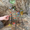 A hand is holding a colorful stick puppet from the Tiger Tribe Nature Art Set by TIGER TRIBE against a tree trunk. Four more puppets, part of this delightful set, lean nearby, adorned with googly eyes, colorful yarn, feathers, and painted spots. Each puppet boasts a whimsical and unique design perfect for creative play for kids.