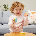 A toddler with light brown hair wearing a light pink shirt is sitting on a wooden table. The child is holding various Tiger Tribe Shape Shakers and smiling. A gray couch adorned with a floral pillow and a plant in the background complete the scene, making it perfect for playful learning with TIGER TRIBE Shape Shakers.