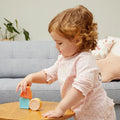 A young child with curly hair, wearing a light pink plaid shirt, is playing with colorful TIGER TRIBE Shape Shakers on a wooden table. A grey sofa and some cushions are visible in the background. The child appears focused on stacking the blocks.