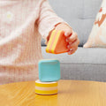 A child wearing a pink and white striped shirt is stacking colorful geometric blocks from the TIGER TRIBE Tiger Tribe Shape Shakers on a wooden table. The bioplastic blocks are orange, blue, and yellow, featuring shapes like circles, a square, and a triangle. A grey couch and a cushion are in the background.