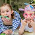 Two children are playfully drawing on a glass surface using TIGER TRIBE Window Markers. The child on the left, wearing a blue-checkered dress, has drawn a green mustache and stick-out tongue. The child on the right, in a white shirt, has added colorful glasses and a propeller hat. Both are making funny faces.