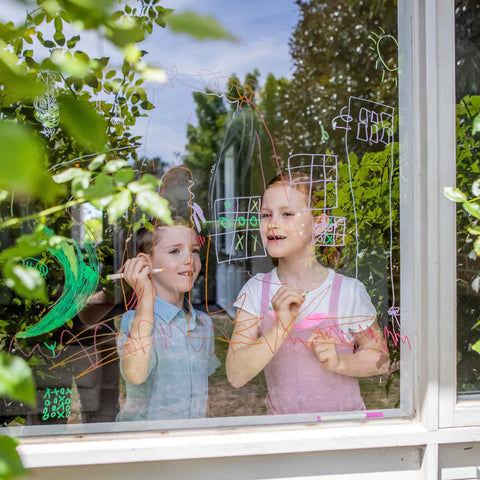 Two children, a boy and a girl, are drawing colorful shapes, patterns, and illustrations using TIGER TRIBE's kid-safe Tiger Tribe Window Markers on a window from the outside. Bright green foliage surrounds the window, and the children look focused and engaged in their artistic activity.