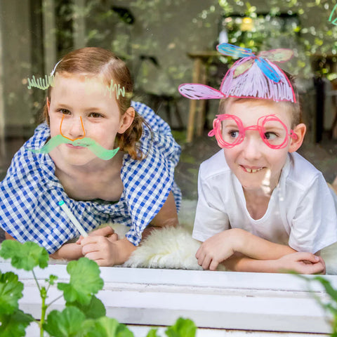 Two children are seen through a window, with playful doodles on the glass aligning with their faces. Using TIGER TRIBE Window Markers, the child on the left has drawn a mustache and curly eyebrows, while the child on the right has created large glasses and colorful hair. They both look cheerful.