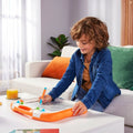 A curly-haired child wearing a denim jacket kneels at a white table, focused on drawing with an orange Tomy Megasketcher. The table also features a glass of orange juice and a book. In the background, a couch adorned with colorful pillows and a plant enhances the cozy ambiance, making it an ideal space for mess-free creativity.